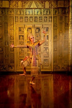Vertical image of Asian man with Thai traditional cloth dance show in front of Thai painting on wall of public place.