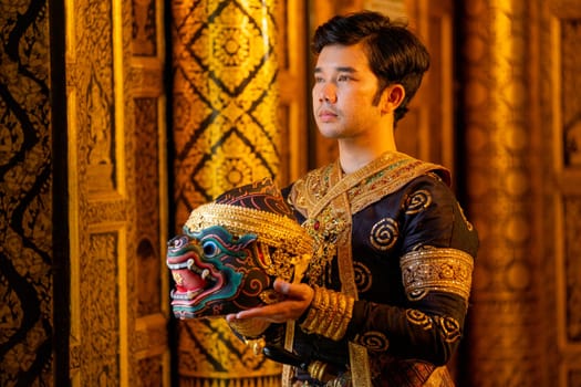 Asian man with traditional cloths relate to Khon or Thai classic masked stand with holding black monkey mask in front of windows of public building also look outside.