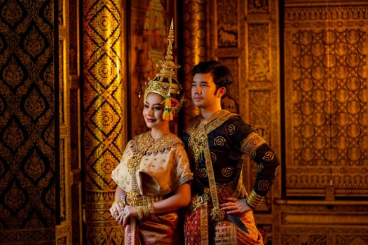 Beautiful Asian woman with Thai traditional dress stand beside Asian man with black traditional dress in front of windows and look outside.