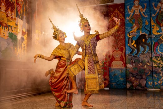 Beautiful Asian woman wear Thai traditional dress action of dancing together with man stand in front of Thai painting on public place wall.