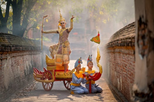 Asian man with old Thai traditional cloth hold weapon and stand and action of dance on traditional chariot also hold weapon stay in front of ancient building.