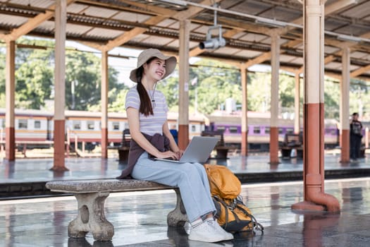 Young Asian woman in modern train station Female backpacker passenger sitting on a bench using a laptop while waiting for a train