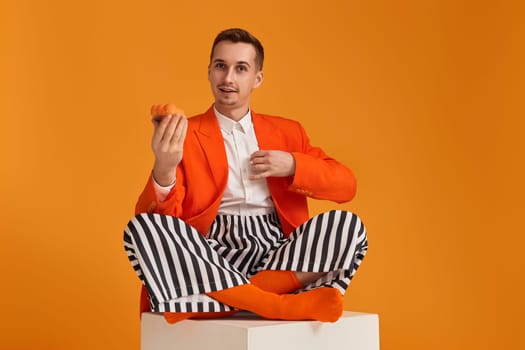 extravagant funny young man in orange jacket and striped pants holding cupcake on orange background.