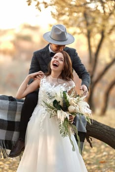 beautiful happy stylish bride and groom in hat laughing near tree in autumn