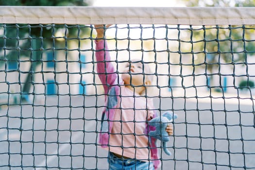 Little girl with a soft toy stands near the tennis net, touching its top with her hand. High quality photo