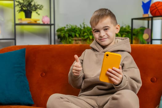Young child boy texting share messages content on smartphone social media applications online watching relax movie. Thumbs up. Caucasian preteen kid uses mobile phone at home in play room sits on sofa