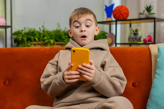 Young child boy texting share messages content on smartphone social media applications online watching relax movie. Amazed shocked Caucasian teenager kid uses mobile phone at home in room sits on sofa