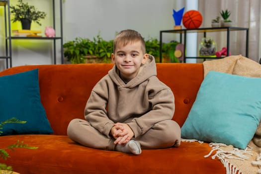 Close-up portrait of happy smiling cheerful Caucasian preteen school boy 8 years old. Young lovely blonde little child kid looking at camera at home play living room apartment sitting on orange couch