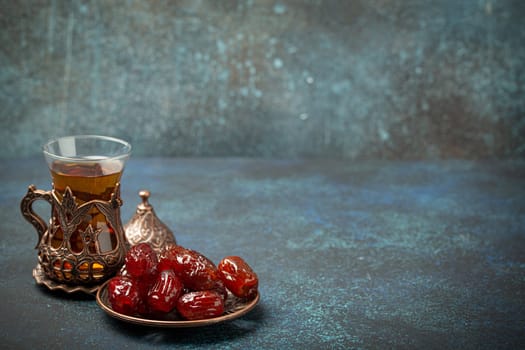 Breaking fasting with dried dates during Ramadan Kareem, Iftar meal with dates and Arab tea in traditional glass, angle view on rustic blue background. Muslim feast, space for text.