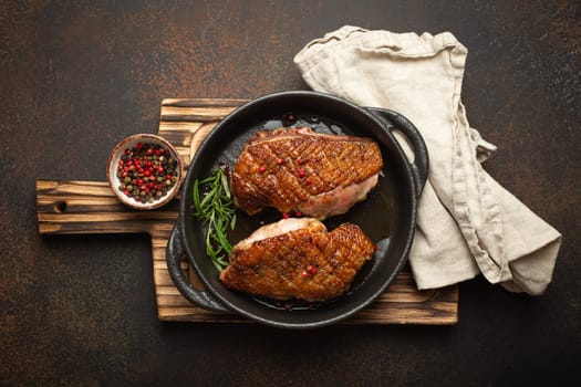 Two roasted duck breast fillets with crispy skin, with pepper and rosemary, top view in black cast iron pan with knife, dark brown concrete rustic background.