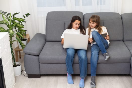 Cute little girl and older sister playing together smiling and having bonding time using a laptop on couch at home. Happy family Siblings relationship and digital technology lifestyle concept. High quality photo