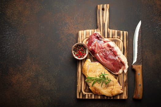 Two raw uncooked duck breast fillets with skin, seasoned with salt, pepper, rosemary top view on wooden cutting board with knife, dark brown concrete rustic background, space for text.