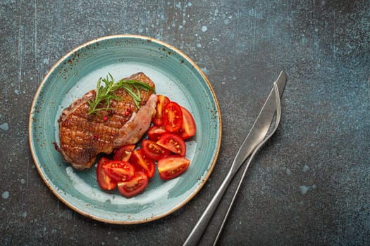 Delicious roasted duck breast fillet with golden crispy skin, with pepper and rosemary, top view on ceramic blue plate served with cherry tomatoes salad, rustic concrete rustic background