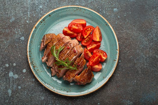 Delicious roasted sliced duck breast fillet with golden crispy skin, with pepper and rosemary, top view on ceramic blue plate served with cherry tomatoes salad, rustic concrete rustic background.
