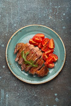 Delicious roasted sliced duck breast fillet with golden crispy skin, with pepper and rosemary, top view on ceramic blue plate served with cherry tomatoes salad, rustic concrete rustic background.