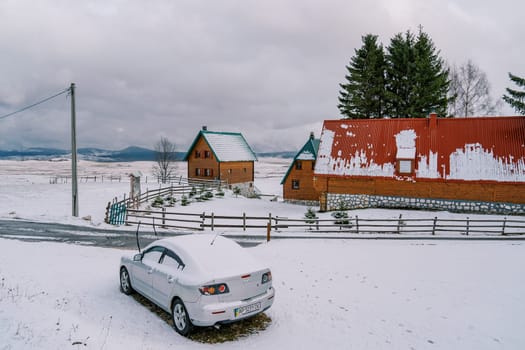 Snow-covered car stands by the road in front of wooden cottages. High quality photo