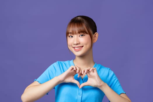 Young asian woman shows her fingers heart-shaped sign and presses to chest on purple background.