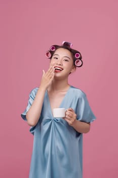 Beautiful young woman in blue dress and hair curlers drinking coffee on pink background
