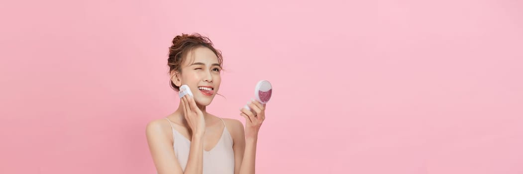  Young attractive lady applying cushion on her face with powder puff on pink background
