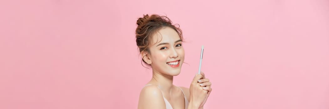 Smiling young asian woman holding blush brush near face isolated on pink