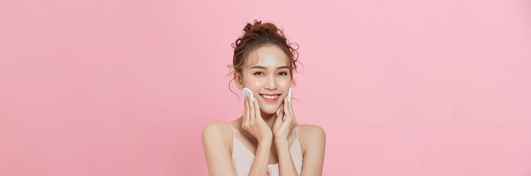 Cheerful young woman holding cotton pad while standing isolated over pink