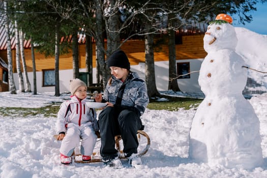 Mom feeds a little girl porridge from a spoon, sitting on a sleigh near a snowman in the yard. High quality photo