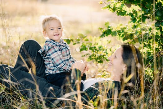 Smiling little girl sitting on belly of mother lying in tall grass. High quality photo