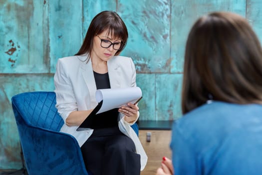 Middle-aged woman counselor psychologist with clipboard sitting in office chair working with patient. Mature female professional mental specialist psychotherapist therapist social worker, workplace