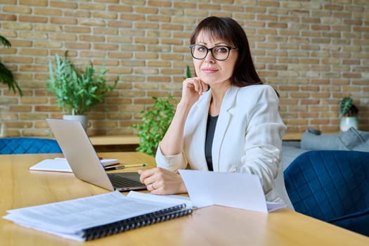 Middle-aged confident business woman office worker, manager looking at camera, sitting at desk with laptop computer. Business, work, job, management, finance, law, logistics, sales, services concept