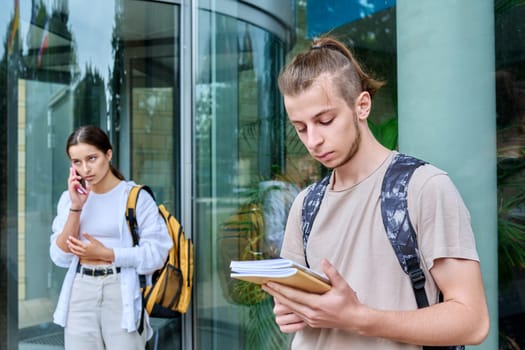 Hipster guy teenager student 18, 19 years old with backpack notebooks standing outdoor, educational building background. Youth, education, lifestyle concept