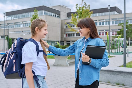 Talking female teacher and schoolgirl child outdoor, school building background. Meeting communication student girl with backpack and mentor counselor. Education, pre-teenage, learning, back to school