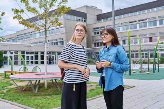 Talking female teacher and teenage high schoolgirl outdoor, school building background. Meeting communication student girl with backpack and mentor counselor. Education, adolescence, learning concept