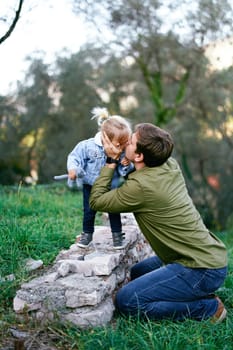 Dad sits on his knees and kisses a little girl standing on a stone fence. High quality photo