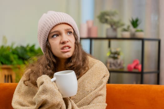 Caucasian sick child girl wear hat wrapped in plaid sit alone shivering from cold on sofa drinking hot tea in unheated home without heating due debt. Unhealthy kid feeling discomfort try to warming up