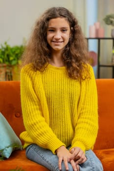 Close-up portrait of happy smiling cheerful Caucasian school girl 15 years old. Young adult lovely child kid student looking at camera at home living room apartment sitting on orange couch. Vertical