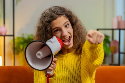 Child girl talking with megaphone, proclaiming news, loudly announcing advertisement warning using loudspeaker to shout speech pointing finger to camera. Teenager kid at home room apartment on sofa