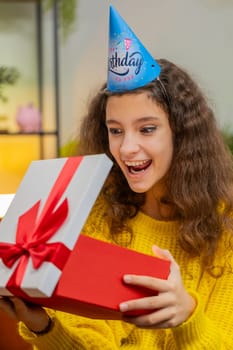 Happy 14-15 years child girl wears festive birthday cap hat hold gift box with ribbon congratulating. Teenager kid celebrating party event opening delivery greeting present at home room. Vertical