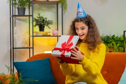 Happy 14-15 years child girl wears festive birthday cap hat hold gift box with ribbon congratulating. Teenager female kid celebrating party event opening delivery greeting present at home room on sofa