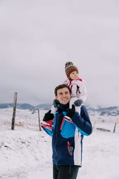 Dad with a little girl on his shoulders walks through a snowy village, looking to the side. High quality photo
