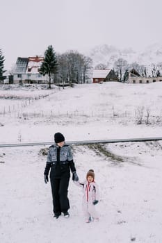 Mom and little girl walk along a snowy hill in the village, holding hands. High quality photo