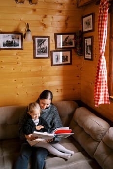 Mom reads a book to a little girl sitting on her lap on a corner sofa in a wooden house. High quality photo
