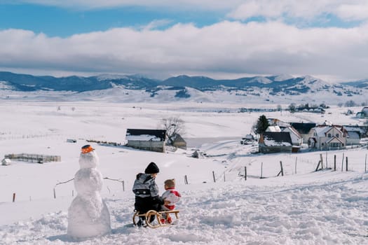 Mom and little girl are sitting on a sled on a snow-capped plain near a snowman and look at each other. Back view. High quality photo