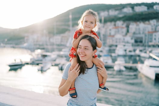 Captured in the warm glow of the setting sun, a smiling mother carries her little daughter on her shoulders. They are enjoying a delightful day at a marina filled with boats