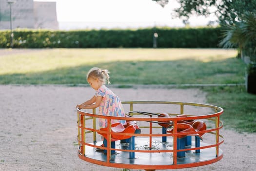 Little girl is standing on a carousel, holding onto a handrail in the park. Side view. High quality photo