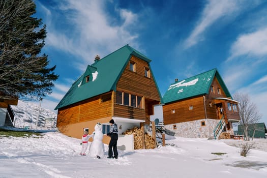 Mom and little girl decorate a snowman near wooden chalets in the snow. High quality photo