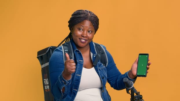 African american bike messenger vertically holding mobile device with isolated green screen display. Female courier showing delivery app on smartphone with chromakey mockup template.