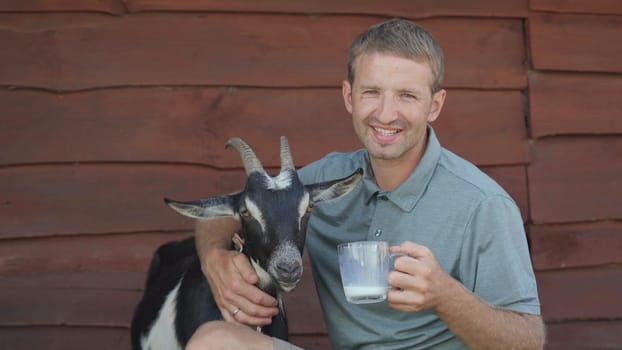 The farmer drinks goat milk from a mug and hugs his beloved goat
