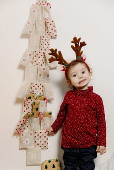 One little Caucasian girl with a headband of deer antlers with a sweet smile on her face takes out a gift from the pocket of a wall advent calendar with her hand, close-up side view.