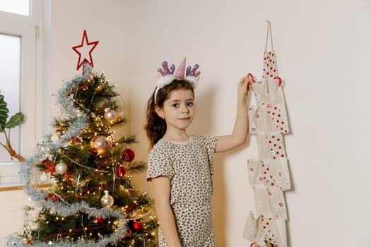One beautiful Caucasian brunette girl with a unicorn horn headband stands by the Christmas tree and with her hand takes out a lollipop from a pocket with the number two of an advent calendar in the morning in the living room, close-up side view.