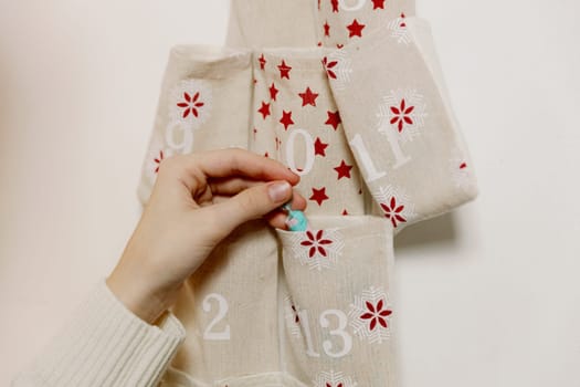 The hand of one Caucasian unrecognizable teenage girl takes out a gift from the pocket number 13 of the advent calendar, hanging on a white wall early in the morning in the living room, close-up side view.